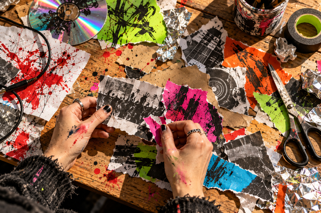 Mesa de trabajo creativa con recortes de papel, pintura, tijeras, cinta, un disco y manos trabajando con materiales de collage.